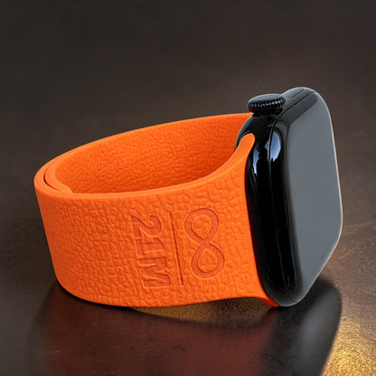 Detailaufnahme eines orangefarbenen Apple Watch Silikonarmbands mit 21M Symbol Prägung, liegend fotografiert